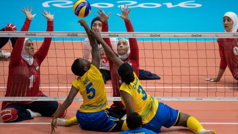 Two women's sitting volleyball teams playing at the Rio 2016 Paralympics. Two female players in yellow uniform are hitting the ball over the net, while two female players in red uniform are trying to block the ball.
