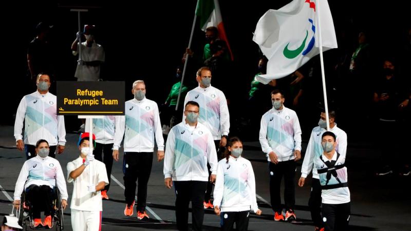 Ten athletes and officials take part in the athletes' parade at the opening ceremony of the Tokyo 2020 Paralympic Games. A volunteer is holding a sign that says "Refugee Paralympic Team" and a male athlete is carrying a flag of the Three Agitos.