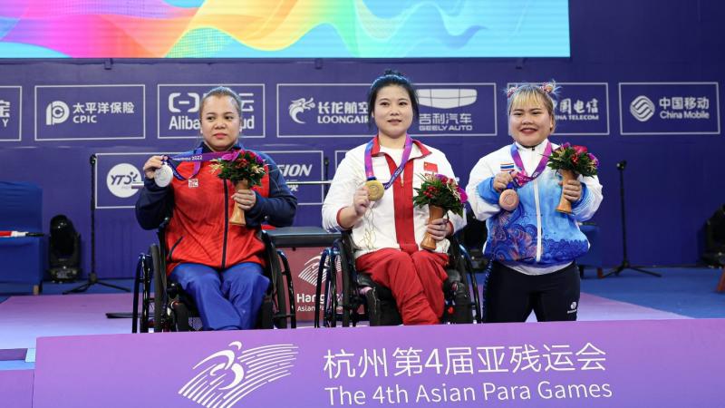 Two women in wheelchairs next to a short stature woman on a podium at the Asian Para Games