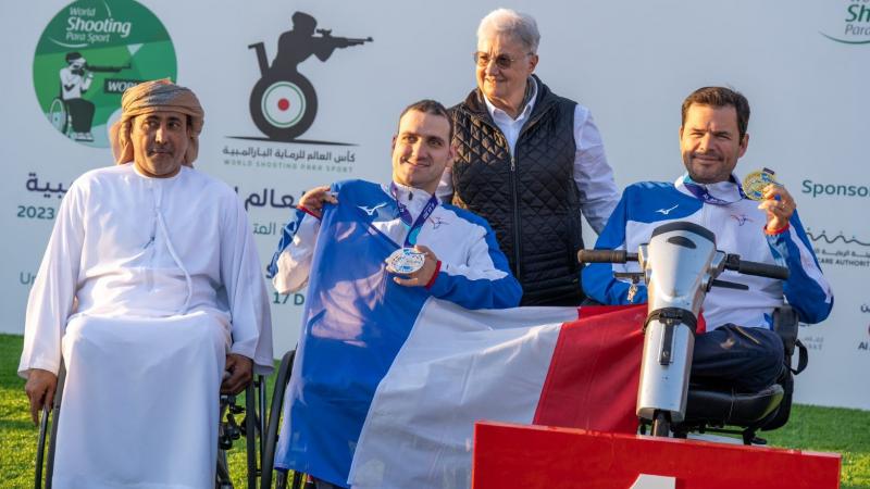 Two men in wheelchairs holding a French flag next to a man in a wheelchair and a woman standing behind them