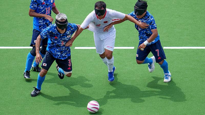 A blind football match between Japan and China at the Tokyo 2020 Paralympics. Three Japanese athletes wearing a blue jersey are trying to defend the ball from a Chinese athlete in a white jersey.