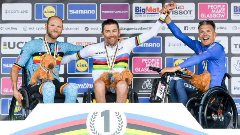 Three male athletes pose for a photo on a podium. The athlete in the centre is wearing the UCI rainbow jersey.