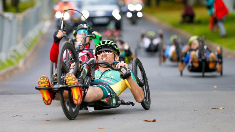 Six athletes compete on handcycles. The athlete leading the pack is wearing a South African jersey.