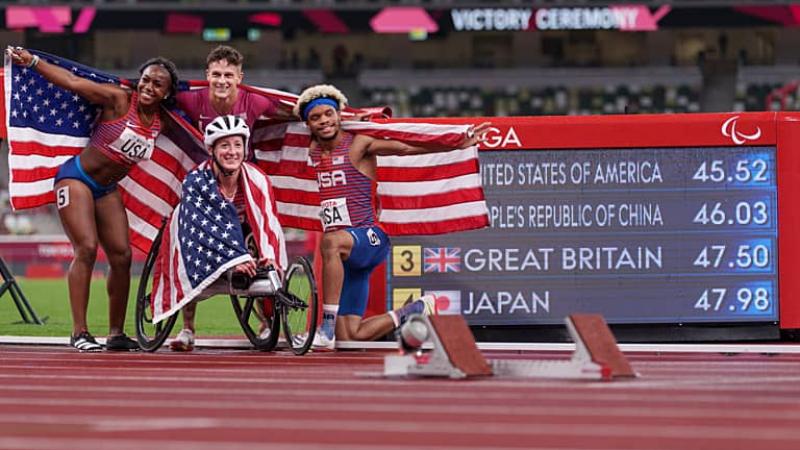 Four US athletes, two women and two men stand next to the scoreboard holding USA flags