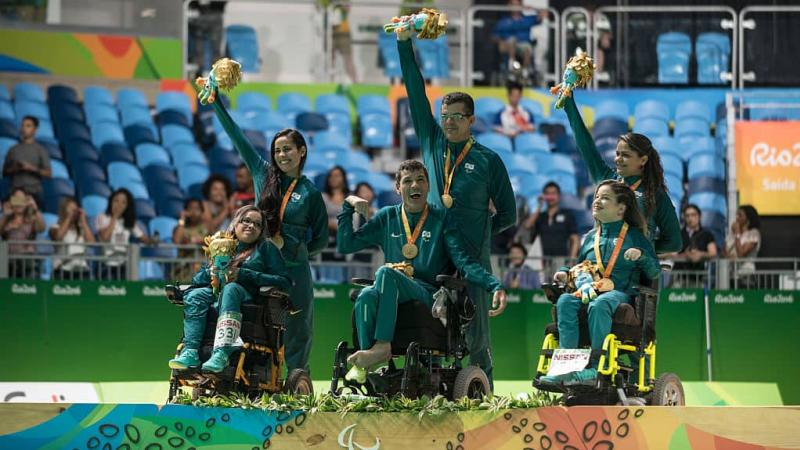 three boccia players in wheelchairs and three standing assistants with their medals on the podium