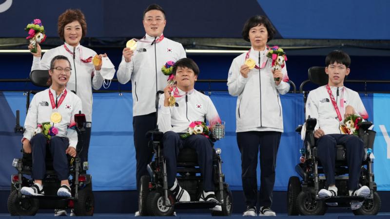 three boccia players in wheelchairs and their standing assistants hold up their gold medals on the podium
