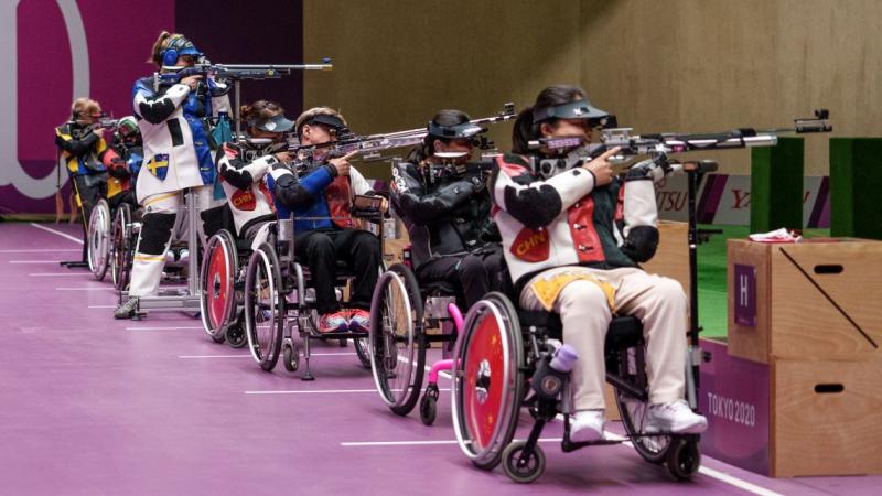 Six female athletes compete in Para shooting competition. Five athletes are competing in a wheelchair, while one athlete is standing.