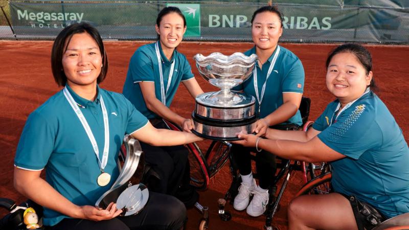 Four female wheelchair tennis athletes pose for a photograph. They are holding a silver trophy.