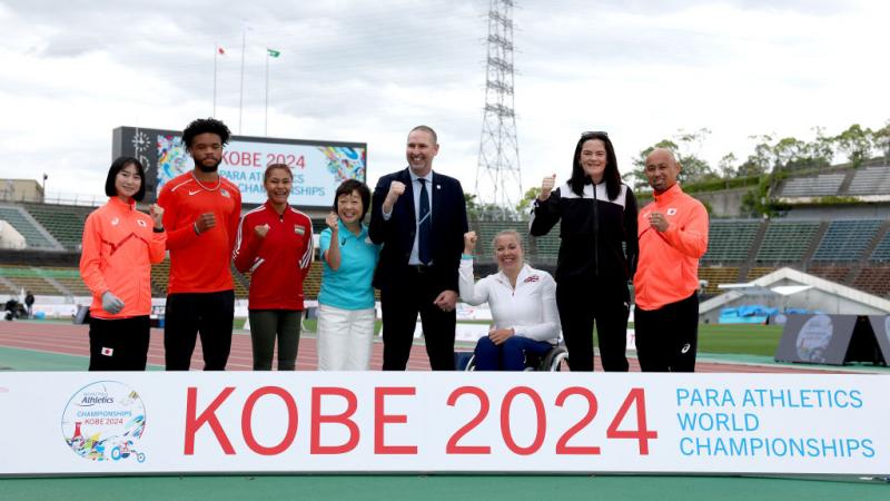 Seven standing person and a wheelchair user posing for a picture in an athletics track