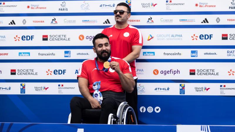 A man in a wheelchair showing a medal with a man standing behind him