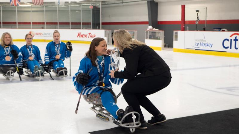 A female Para ice hockey player receiving a trophy from a woman in a hockey ring