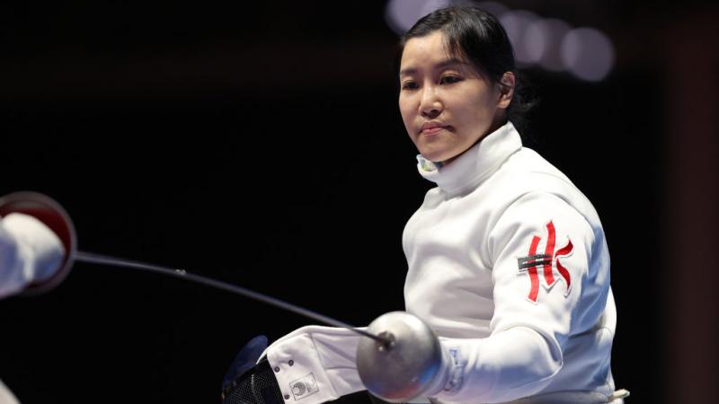 A female wheelchair fencer strikes a pose. She is holding her sword with her left and her mask with her right