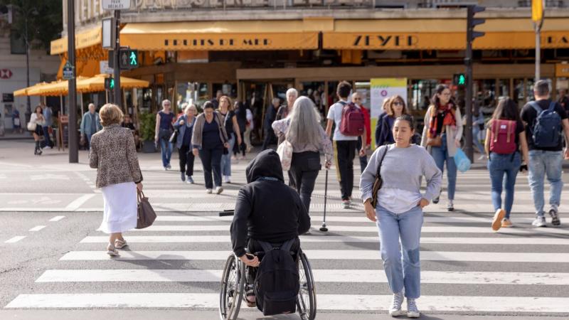 Many people cross a sidewalk in Paris. There are people in a wheelchair and people using canes.