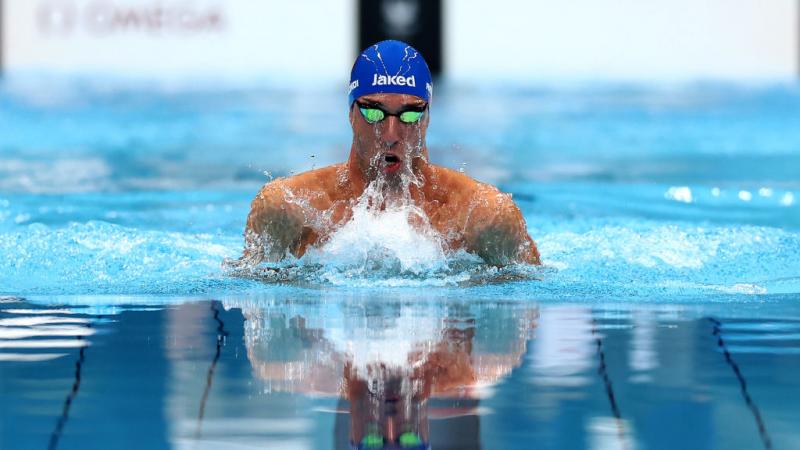 The head and shoulders of a swimmer in a blue cap and googles emerges from the water with a splash