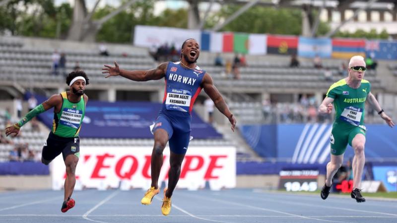Three men cross the finish line on an athletics track. The man in the middle screams and has his arms outstretched.