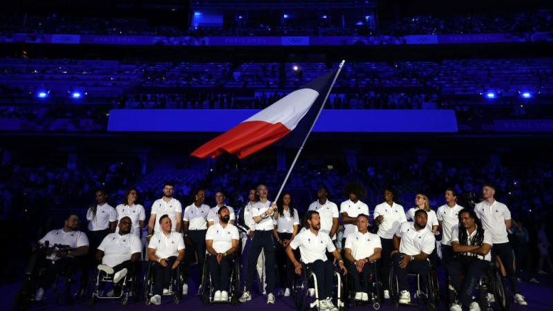 About 20 Paralympic athletes taking part in the Closing Ceremony of the Olympic Games. The female athlete in the centre is waving the French flag.