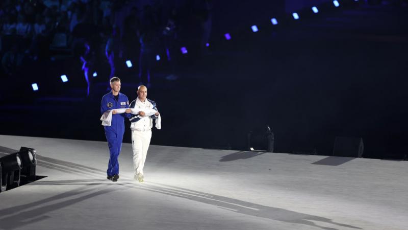 Two men carry a rolled up Paralympic flag across a stage at the Opening Ceremony