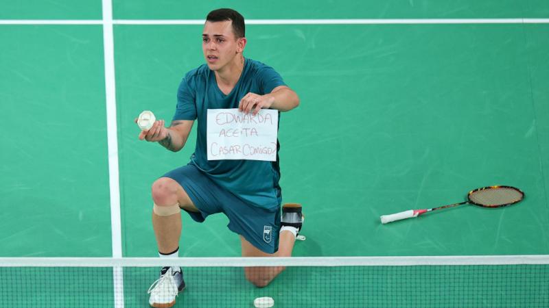 Man in blue tee and shorts kneels on the badminton court holding a ring