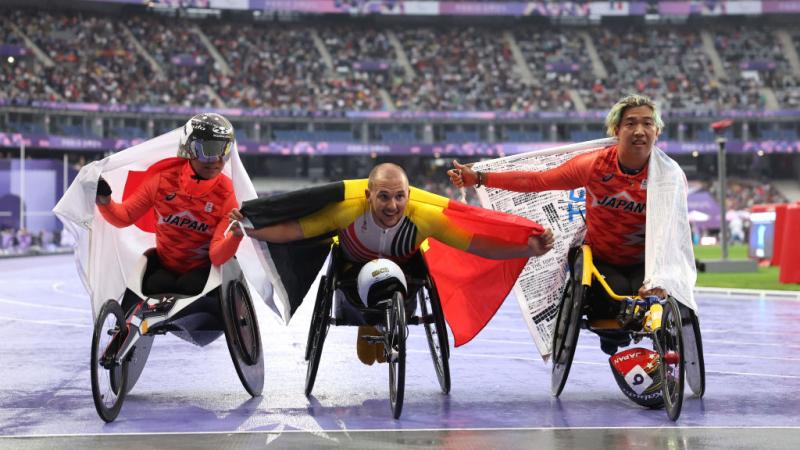 Three athletes in wheelchairs pose for the camera with the flags of their countries (Japan and Belgium) draped around their shoulders
