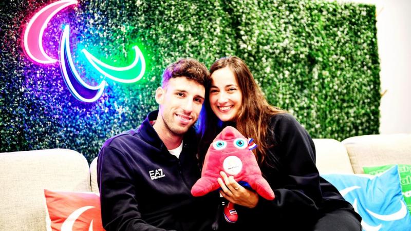 Man and woman face camera dressed in blue with Agitos in neon lights behind them holding the Paris 2024 mascot