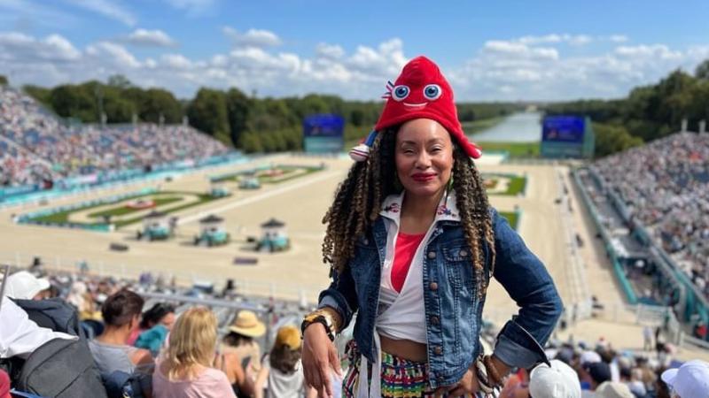 Woman stands facing camera with backdrop of Chateau Versailles Para equestrian wearing Phryge mascot hat