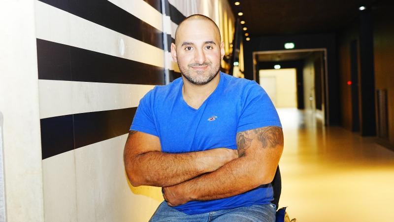 Man seated in blue t-shirt smiles to camera with arms folded
