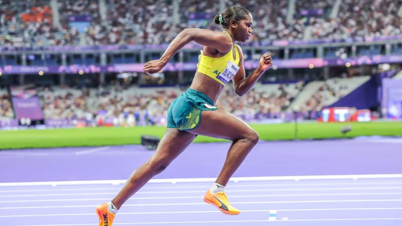 An Australian female athlete with a yellow vest and green shorts runs at the Stade de France athletics stadium