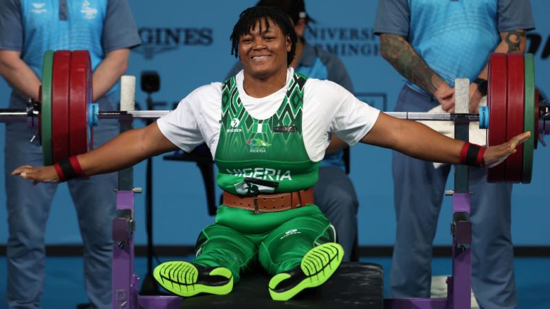 A woman in a green Team Nigeria uniform sits up on a Para powerlifting bench and stretches her arms out wide, smiling