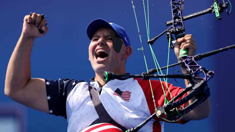 A man raises both arms in celebration, holding his archery bow in one hand