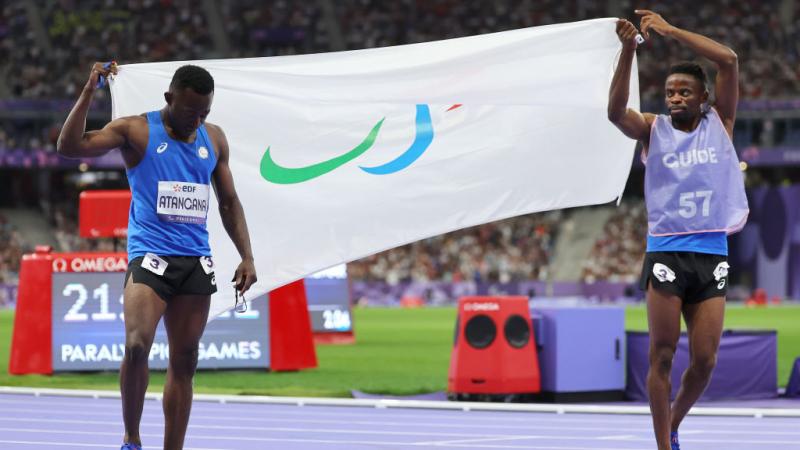 Two sprinters carry opposite sides of a white flag that has the Paralympic Agitos on it