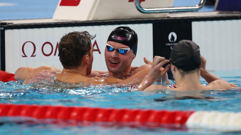 Three men are in a swimming pool, two are congratulating a third, who is wearing a swim cap and goggles.