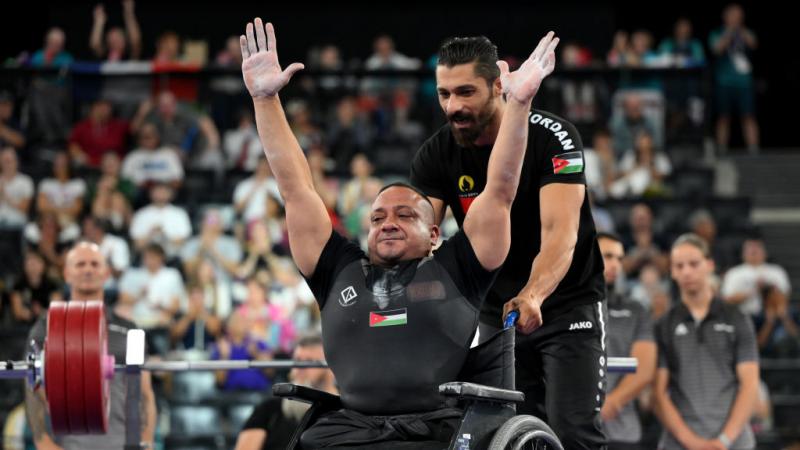 Man in black Jordan kit in wheelchair raises both hands in the air in a sign of celebration while pushed by another man with a beard