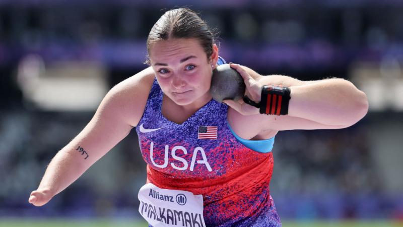 A woman in a Team USA uniform holds a shot put up by her neck to prepare to throw it