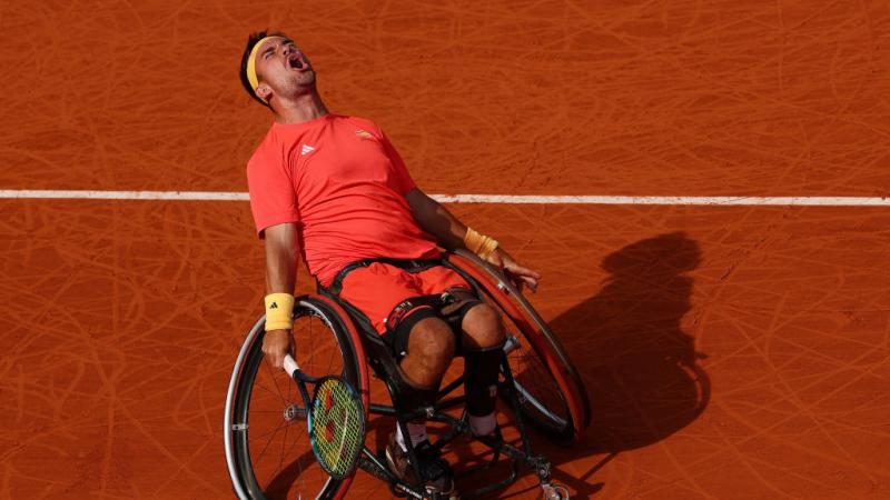 Man leans back in his wheelchair holding a tennis racket closing eyes and screaming, against a backdrop of a clay tennis court
