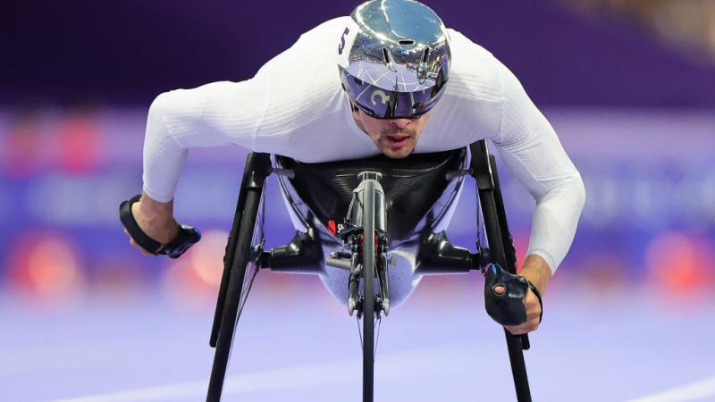 A man in a racing wheelchair and a silver helmet competes on a purple track