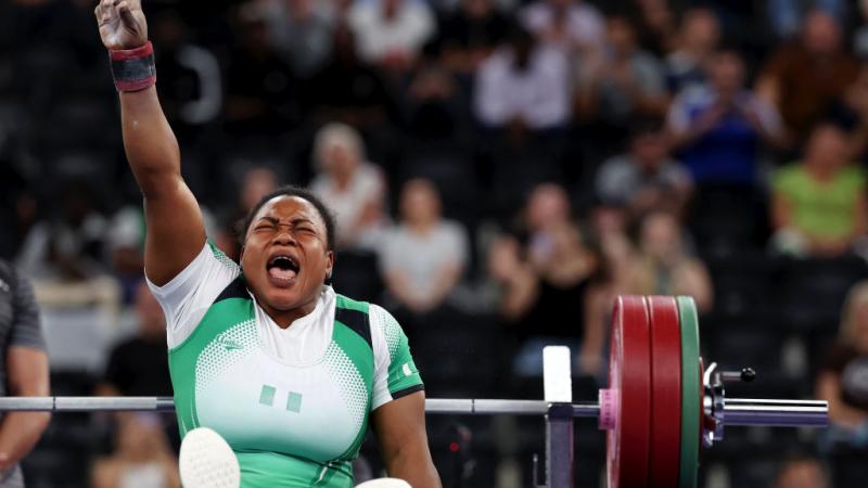 Woman in white and green sport outfit is seated with her hand in the air in a Number 1 sign, she scrunches her face to scream, with powerlifting weights behind her