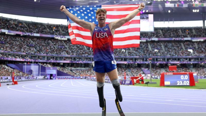 A man with running blades wearing a Team USA uniform holds up an American flag and smiles inside a full stadium