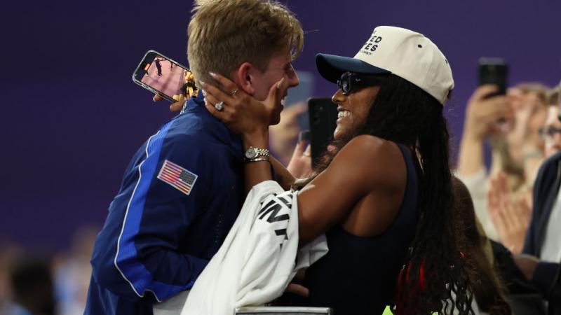 A woman in the crowd in a United States baseball hat puts her arms around a man in a Team USA jacket, both smiling