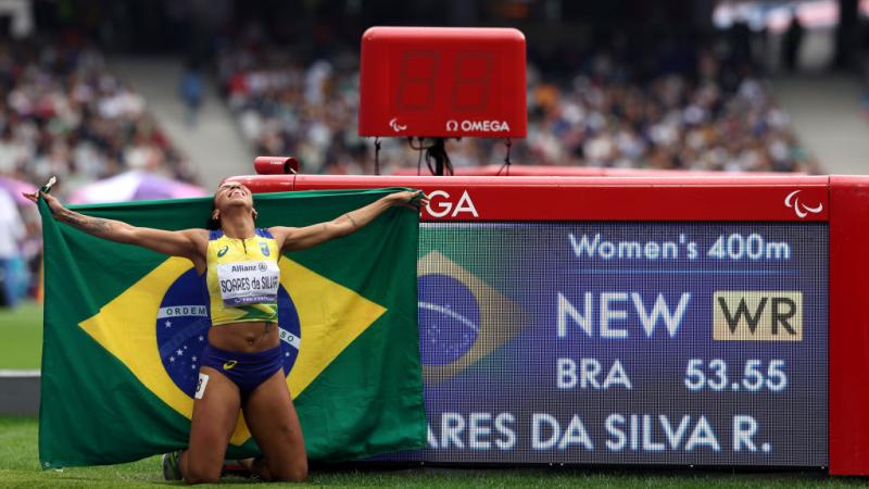A female Brazilian athlete celebrates with her country's flag in front of her new world record of 53.55 on the screen