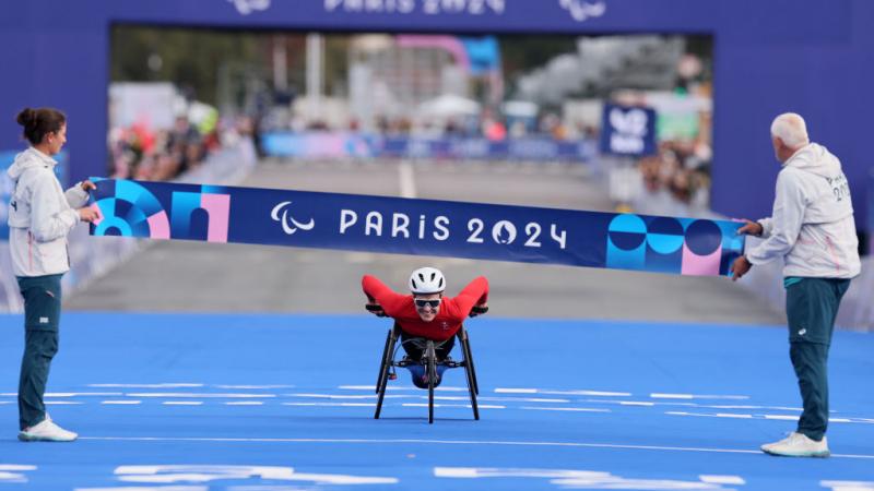 Woman in white helmet, sunglasses and red outfit leans forward in her racing wheelchair with a big smile as she approaches the finish line