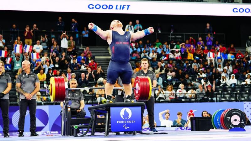 A short stature athlete standing on the bench press in a crowded arena