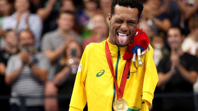 A male Para swimming athlete poses for a photo on the podium. He sticks his tongue 