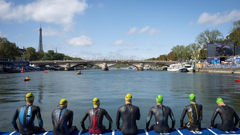 Six Para triathlon athletes are preparing for the swimming segment. The photo captures their back, sitting in front of a river
