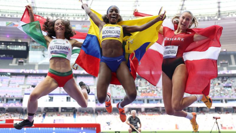 Three female athletes hold their flags and jump to celebrate 