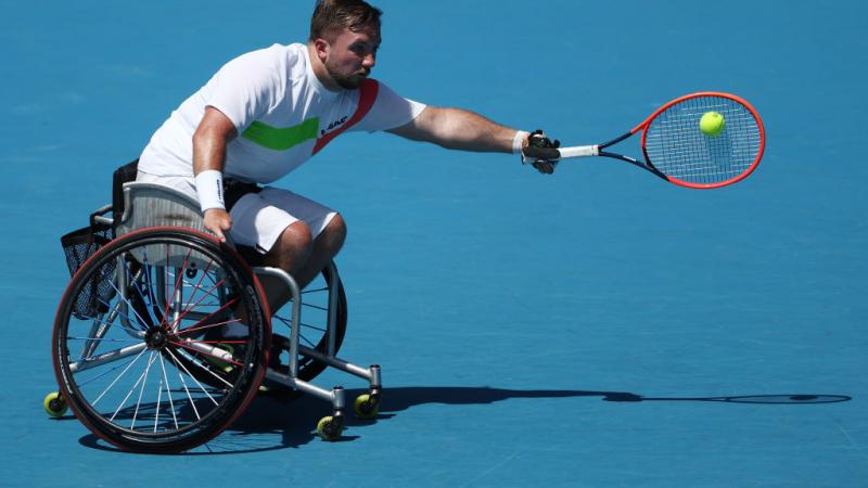 A male wheelchair tennis player reaches to return a shot
