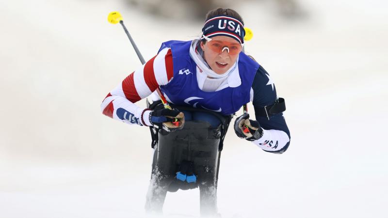 A female athlete competes in a sit-ski. She is wearing a head band that says USA and uniform in red, white and blue.