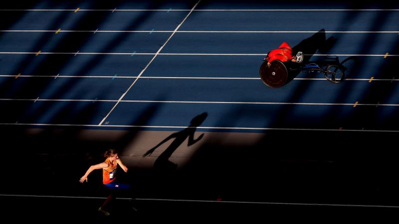 A female athlete with prosthetic legs jumping next to a wheelchair racer in the track