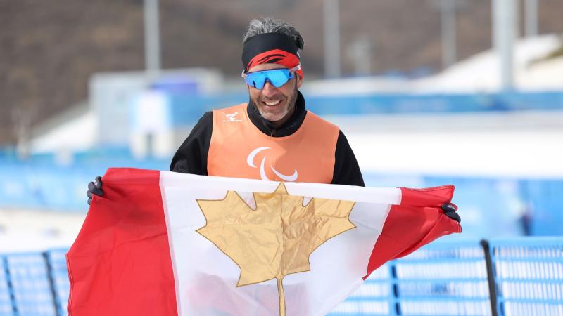 A male athlete wearing sunglasses and an Orange bib holds the Canadian flag