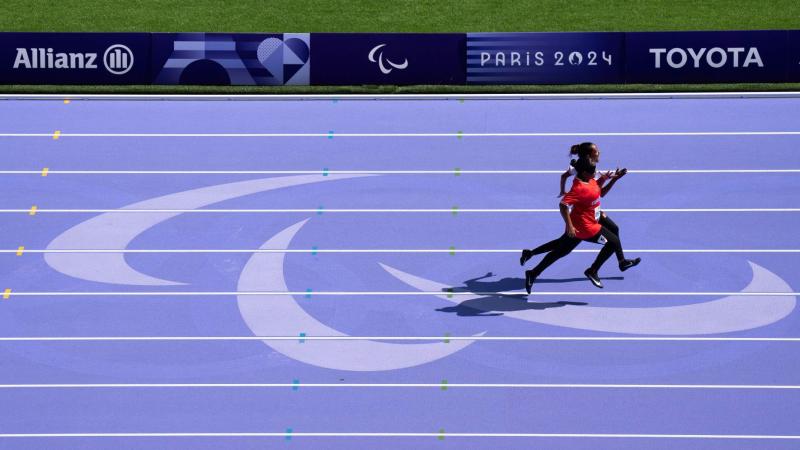 The purple track of the Paris 2024 Paralympics. There is a female athlete competing with her sighted guide