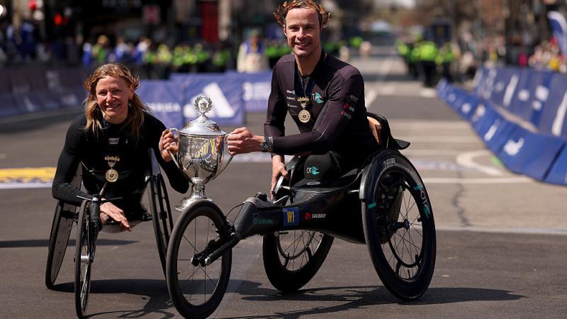 A female and a male wheelchair racer holding the Boston Marathon trophy ahead of the finishing line